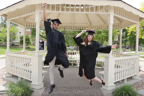 Two students wearing graduation caps and gowns jump in front of the WSU Gazebo.