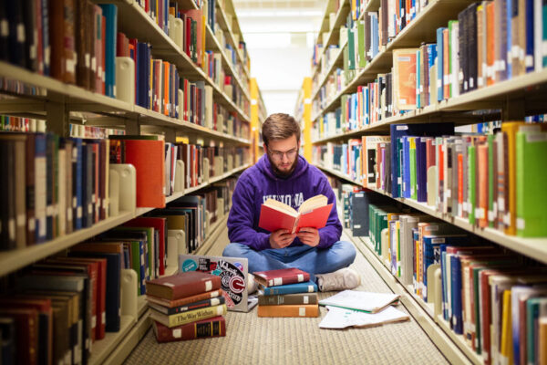 A male WSU student reads a book between the shelves in the library.